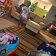 Devon is registered to the contest to win money with this photo: child, standing, living_room, couch, ottoman, rug, plants, houseplants, indoor, portrait, smiling, clothing, t_shirt, shorts, socks, toys, laundry, shopping_bag, home_decor, cozy