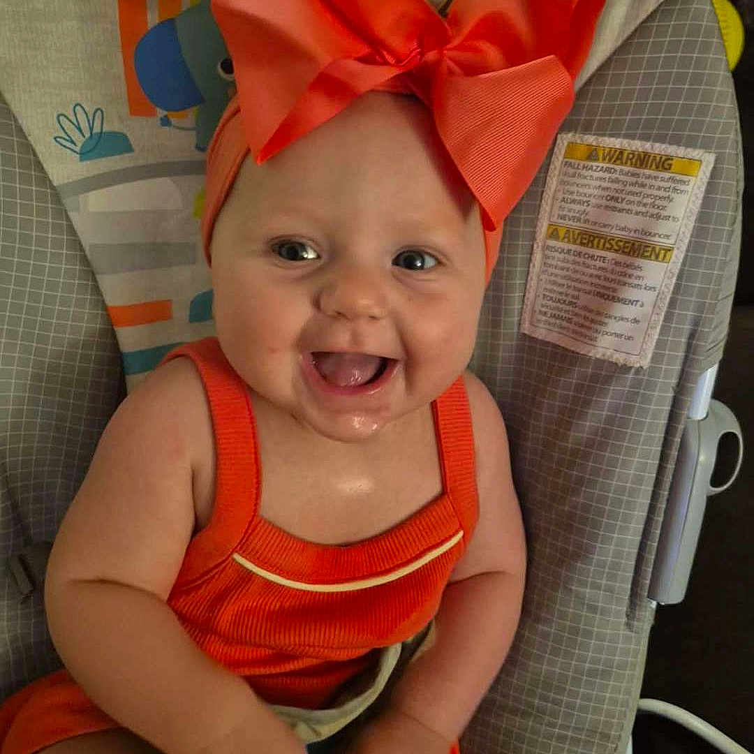 Anara is registered to the contest to win money with this photo: baby, child, smiling, headband, red_bow, orange_outfit, baby_seat, colorful, happy, cute, infant, indoors, person, seat, clothing, cheerful, face, portrait, sitting, playful