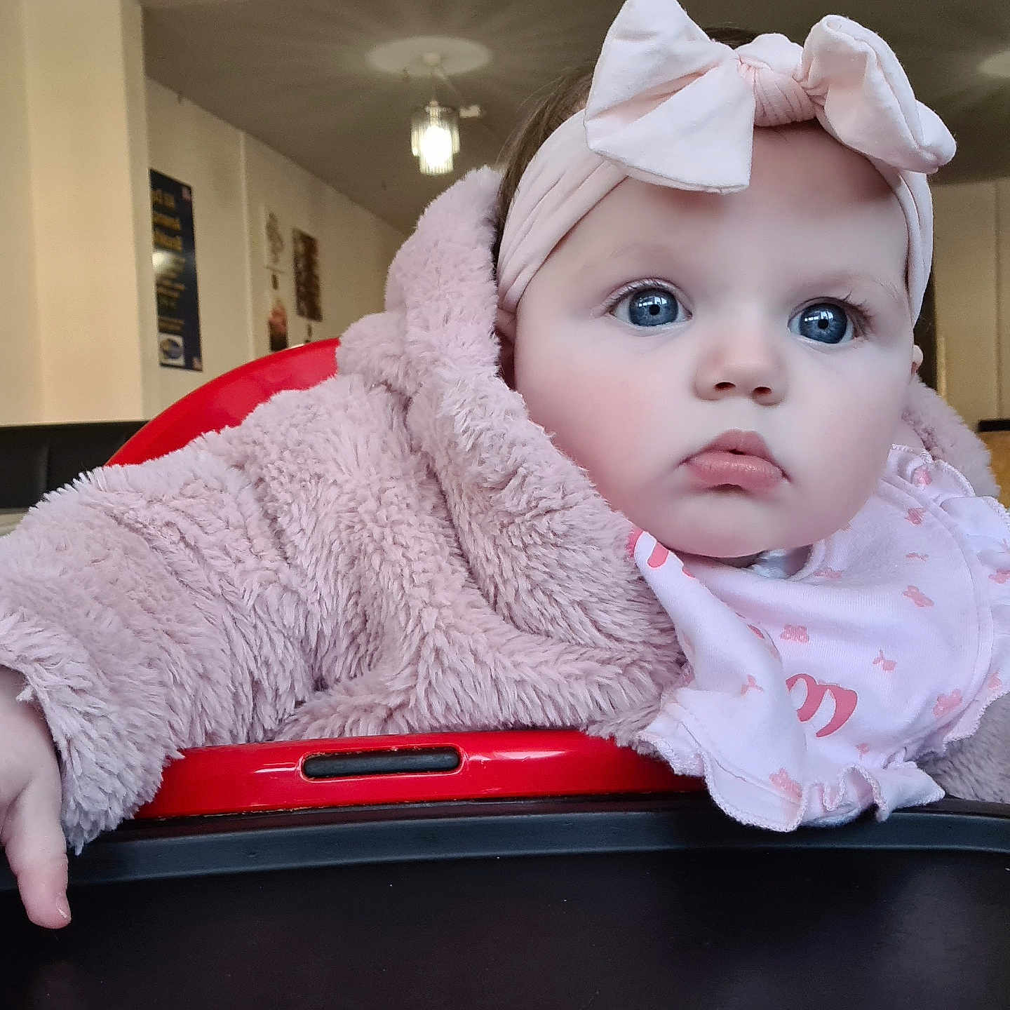 Peyton is registered to the contest to win money with this photo: baby, babygirl, blue_eyes, child, clothing, curious, cute, face, hand, headband, high_chair, indoor, infant, person, pink_jacket, portrait, restaurant, seat, soft_texture, table