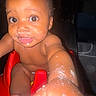 baby, infant, child, sitting, red_seat, reaching, curious, wide_eyes, smiling, face, bare_skin, food, messy, camera_flash, indoor, portrait, close_up, dark_background, arms, legs