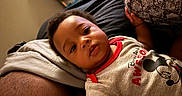 Hezekiah joined the competition — help win amazing prizes! baby, child, infant, portrait, face, lying_down, couch, pillow, indoor, home, clothing, shirt, mickey_mouse, cartoon, black_hair, brown_skin, looking_at_camera, relaxed, close_up, photography