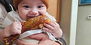 Jaxon is registered to the contest to win money with this photo: baby, child, infant, hat, knit_hat, bear_ears, food, meat, roasted_meat, hands, adult, kitchen, cabinet, smiling, happy, person, indoor, portrait, cute, holding