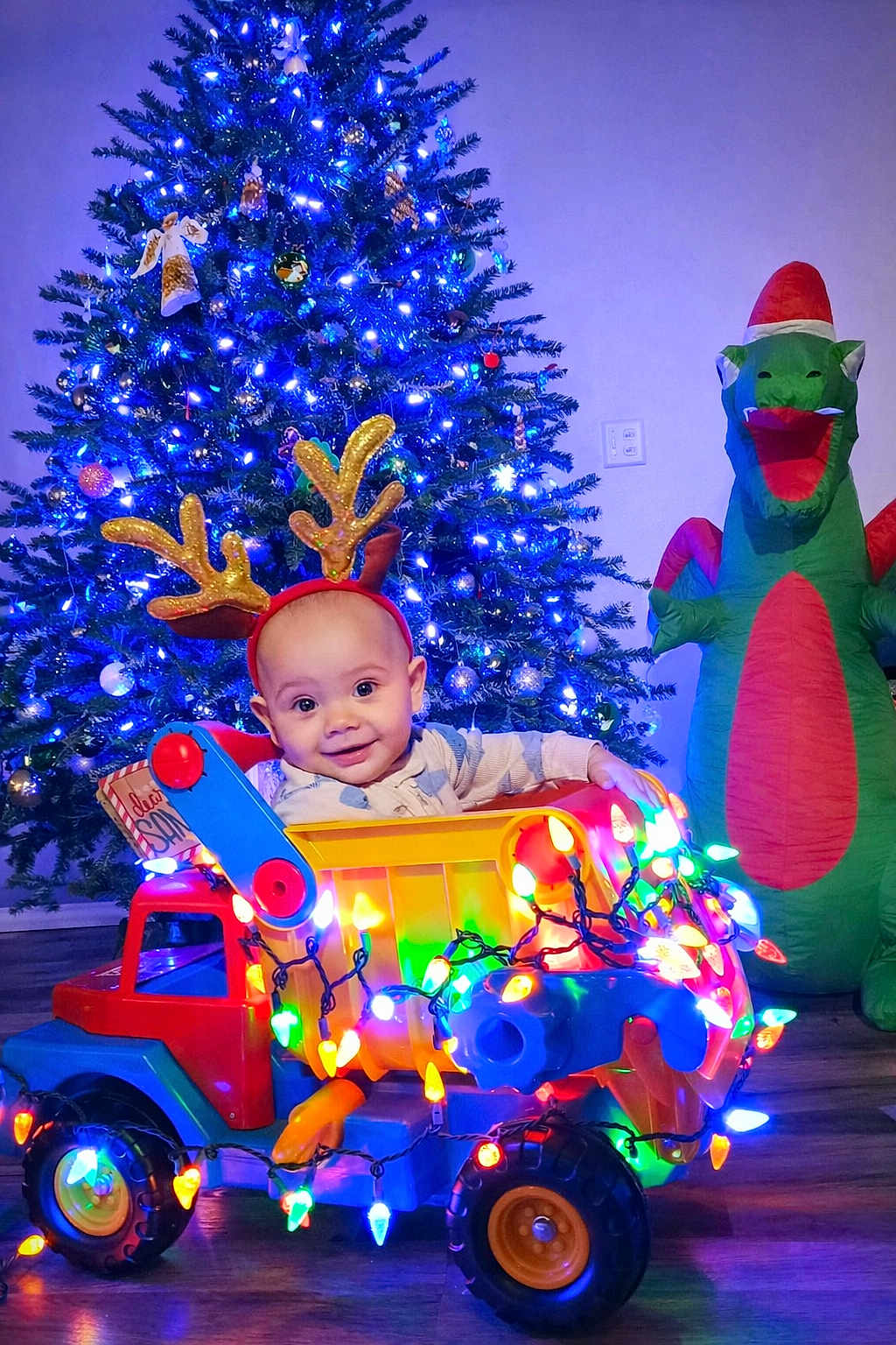 Jaxon is registered to the contest to win money with this photo: baby, reindeer_ears, toy_truck, christmas_lights, christmas_tree, inflatable_dragon, holiday, festive, smiling, indoor, flooring, decorations, colorful, child, playful, seasonal, bright, cute, happy, celebration