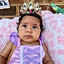 baby, child, crown, princess_dress, purple_dress, pink_blanket, brick_wall, toy, chameleon_toy, wide_eyes, portrait, indoor, cute, costume, headwear, infant, expression, laying_down, soft_texture, decor