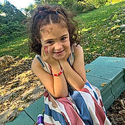 Sophia is registered to the contest to win money with this photo: child, girl, curly_hair, face_paint, bracelet, striped_dress, outdoor, bench, greenery, smile, hands_on_face, nature, sunlight, leaf_litter, playful, portrait, happy, casual, daylight, young