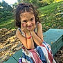 child, girl, curly_hair, face_paint, bracelet, striped_dress, outdoor, bench, greenery, smile, hands_on_face, nature, sunlight, leaf_litter, playful, portrait, happy, casual, daylight, young