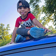 Matteo is registered to the contest to win money with this photo: child, boy, cap, tongue_out, blue_shoes, red_shirt, car_roof, outdoor, trees, nature, playful, smiling, summer, casual_clothing, daylight, young_kid, fun, greenery, sky, park