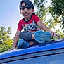 child, boy, cap, tongue_out, blue_shoes, red_shirt, car_roof, outdoor, trees, nature, playful, smiling, summer, casual_clothing, daylight, young_kid, fun, greenery, sky, park