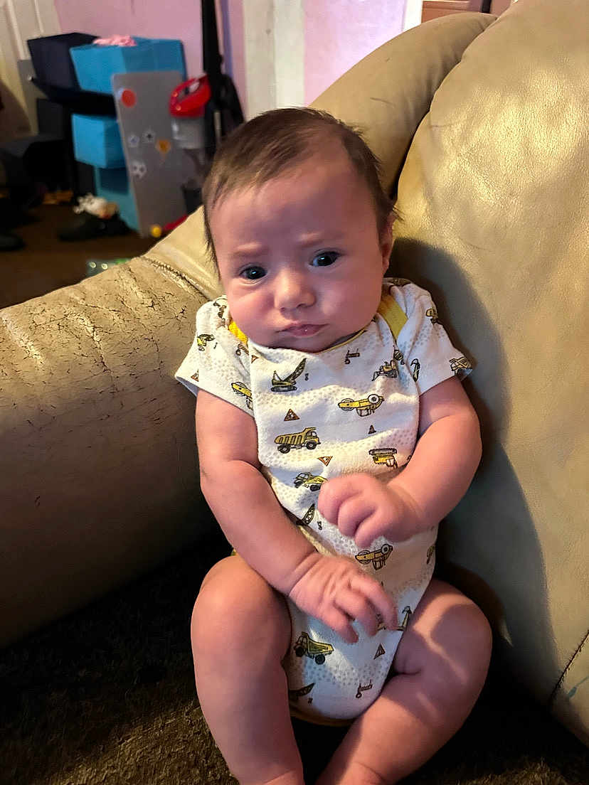 Jamison joined the competition — help win amazing prizes! baby, infant, onesie, construction_theme, chair, leather, indoor, person, expression, clothing, face, sitting, young_child, furniture, home, curious, skin, legs, arms, background