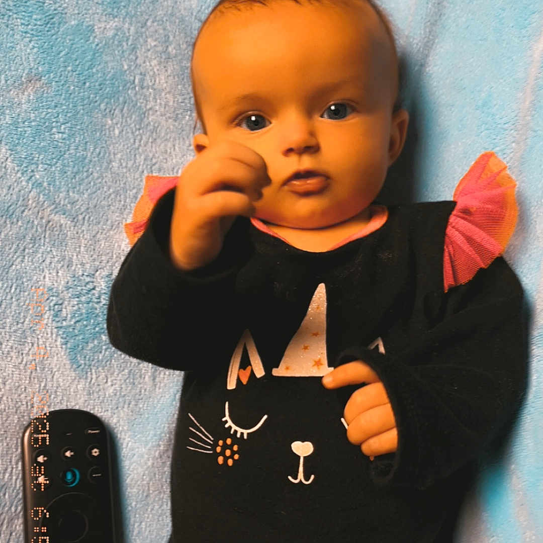 Charlotte is registered to the contest to win money with this photo: baby, black_clothing, blue_blanket, cat_design, child, curious, cute, eyes, face, hand, indoor, infant, lying_down, onesie, party_hat, person, pink_ruffles, remote_control, soft_texture, toddler