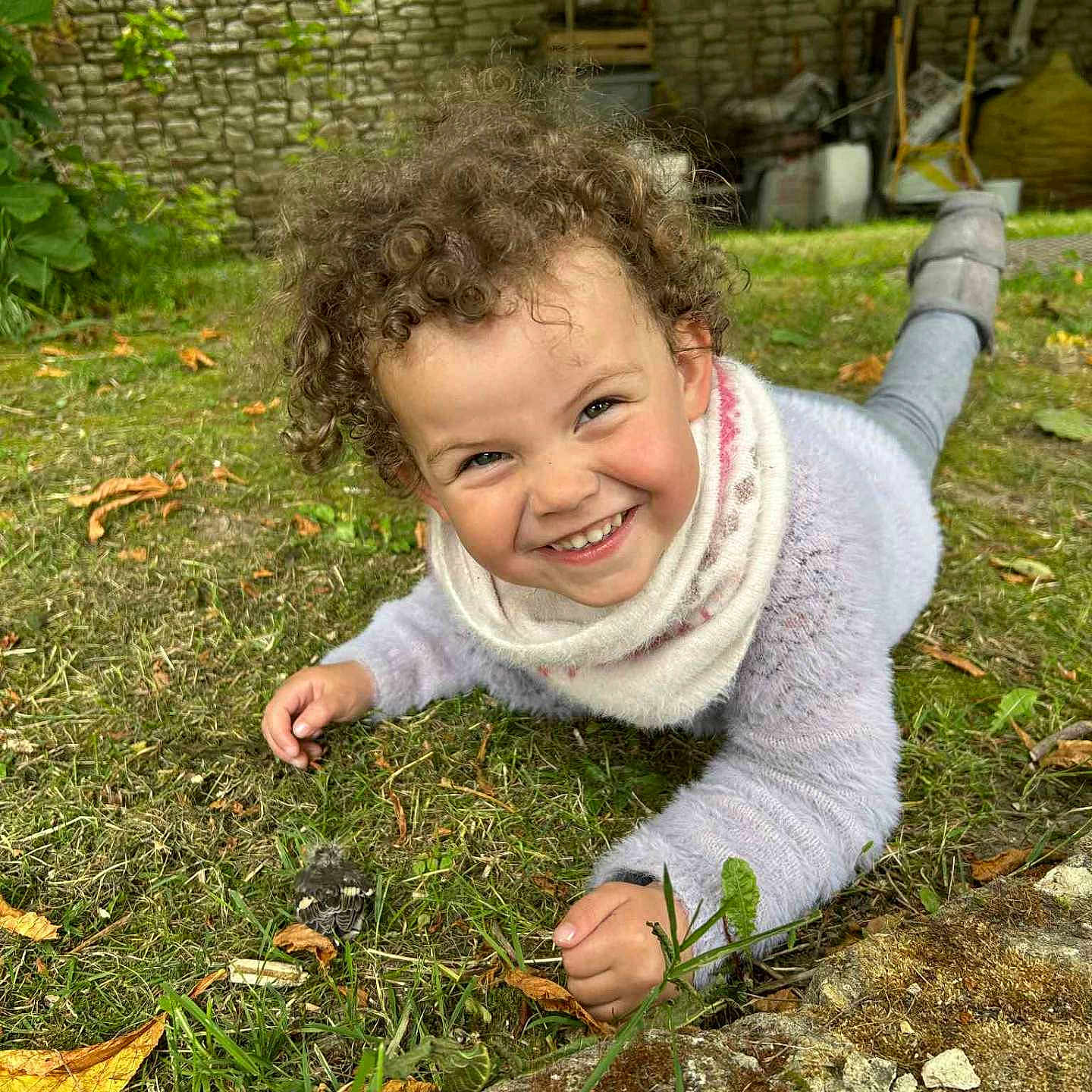 Andréa a rejoint le concours — aidez-le/la à gagner de superbes lots ! casual_clothing, child, curly_hair, daylight, fall, fun, grass, greenery, happy, leaves, nature, outdoor, person, playful, scarf, smile, stone_wall, sweater, toddler, young_child