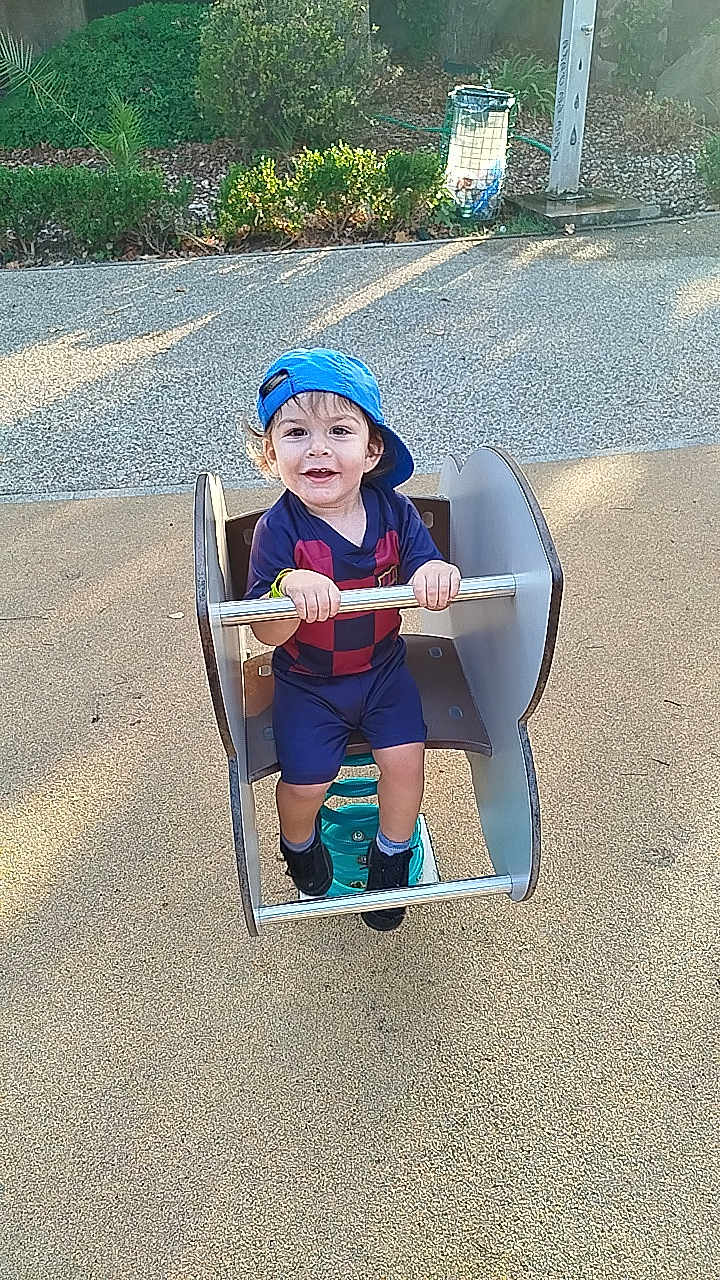 Andrès participe au concours pour gagner de l'argent avec cette photo : child, toddler, playground, spring_rocker, blue_cap, smiling, outdoor, daylight, park, greenery, pavement, casual_clothing, shorts, shoes, metal_bars, happy, fun, young, person, recreation