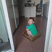 Andrès participe au concours pour gagner de l'argent avec cette photo : toddler, child, green_shirt, cardboard_box, blue_ball, smiling, kitchen, floor, tile_floor, indoor, cabinet, appliance, door, person, happy, young_child, toy, playful, household, sitting