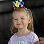 child, toddler, birthday, crown, smiling, happy, celebration, face, person, girl, young_child, casual_clothing, hand, indoor, portrait, light_purple_shirt, short_sleeves, blonde_hair, pigtails, cute