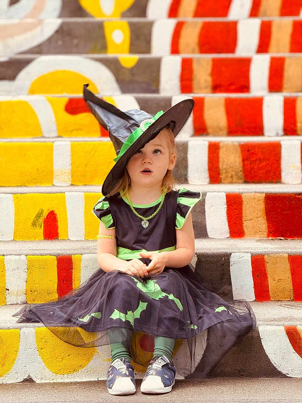 Liza is registered to the contest to win money with this photo: child, costume, witch_hat, dress, stairs, painted_stairs, colorful, outdoor, sitting, curious_expression, green, black, bat_pattern, shoes, casual_shoes, young_child, portrait, holiday, halloween, urban_art