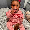 baby, bed, blanket, bow, child, cozy, curly_hair, cute, face, happy, hearts, indoors, patterned_blanket, person, pink_outfit, sitting, smiling, teddy_bear, toddler, toy