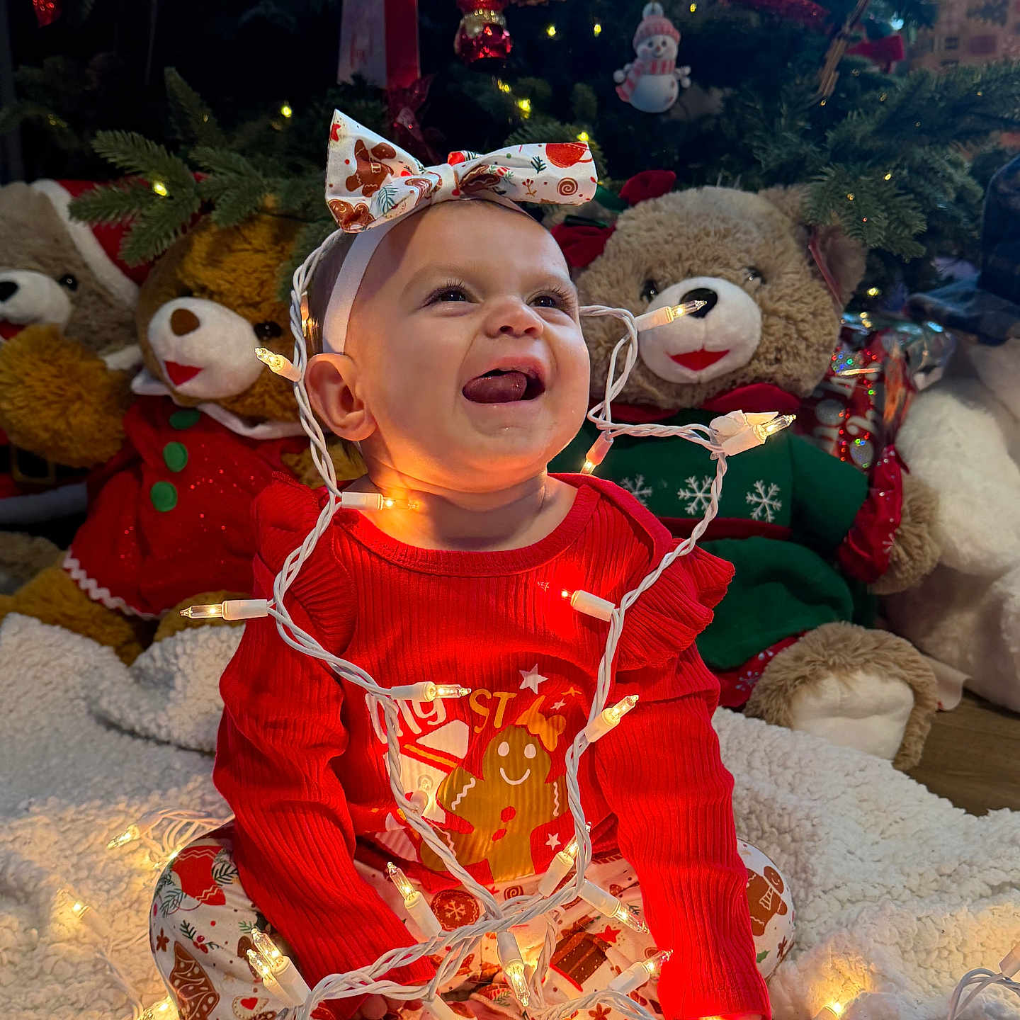 Ellehana is registered to the contest to win money with this photo: baby, blanket, child, christmas_lights, christmas_tree, cute, decorations, festive_clothing, headband, holiday_decor, holiday_season, indoors, joyful, pajamas, plush_toys, sitting, smiling, teddy_bears, tongue_out, warm_lighting