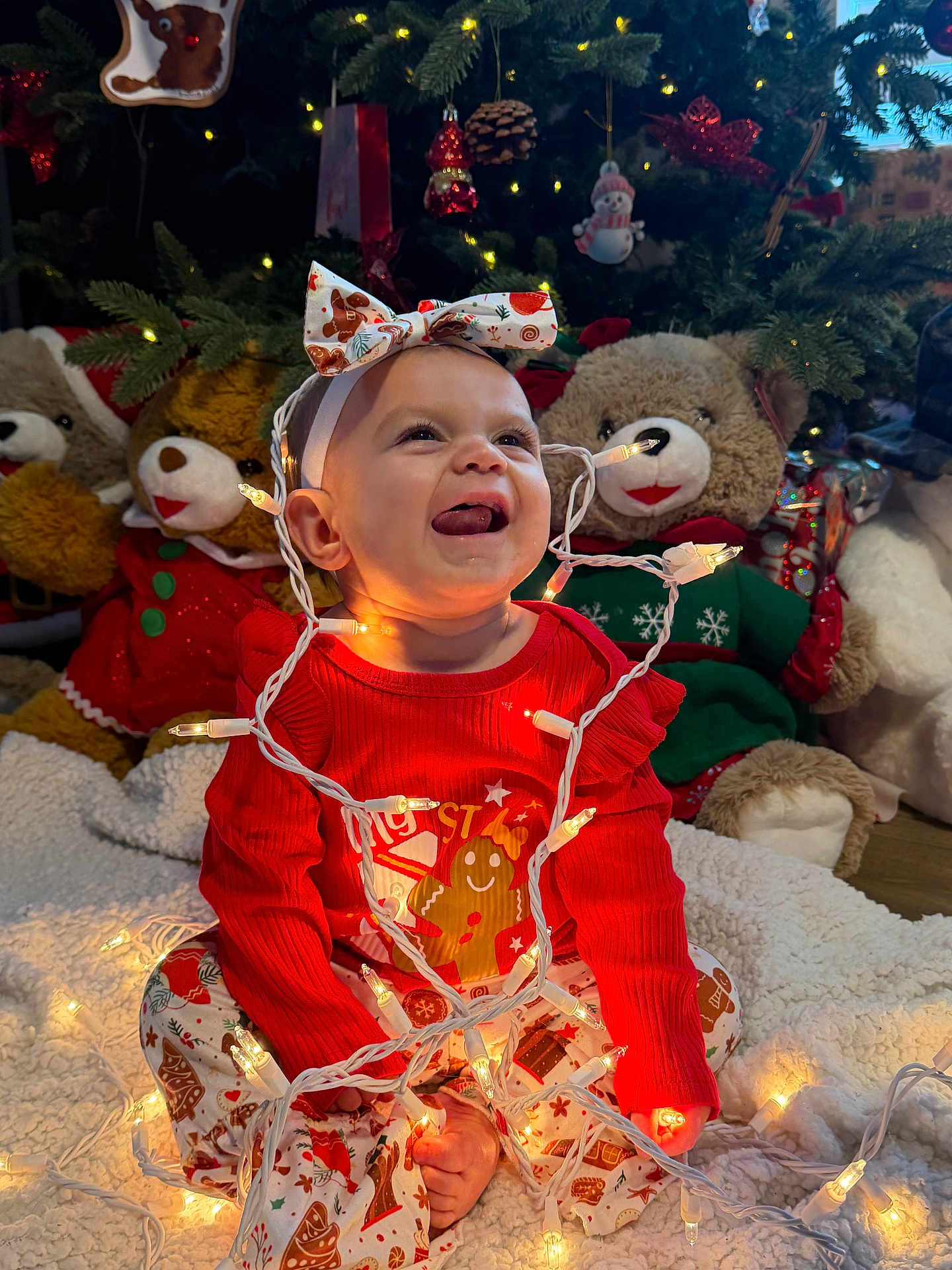 Ellehana is registered to the contest to win money with this photo: baby, blanket, child, christmas_lights, christmas_tree, cute, decorations, festive_clothing, headband, holiday_decor, holiday_season, indoors, joyful, pajamas, plush_toys, sitting, smiling, teddy_bears, tongue_out, warm_lighting