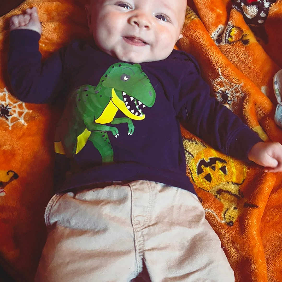 Karsyn joined the competition — help win amazing prizes! baby, child, infant, smiling, happy, portrait, lying_down, blanket, orange_blanket, dinosaur_shirt, t_rex, clothing, long_sleeves, pants, indoors, close_up, face, eyes, playful, cozy