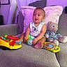 baby, child, toy, teddy_bear, couch, pillow, indoor, colorful, purple_light, smiling, cute, plush, clothing, furniture, person, sitting, playful, infant, home, decor