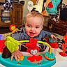 baby, child, smiling, toy, activity_center, indoor, furniture, wooden_cabinet, television, colorful, play, happy, clothing, hand, face, head, room, living_room, elephant_toy, lion_toy