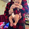 Vyla is registered to the contest to win money with this photo: baby, child, person, adult, holding, smile, indoor, plaid_shirt, red, black, living_room, sofa, clothing, furniture, carpet, window_blinds, toy, pacifier, home, family