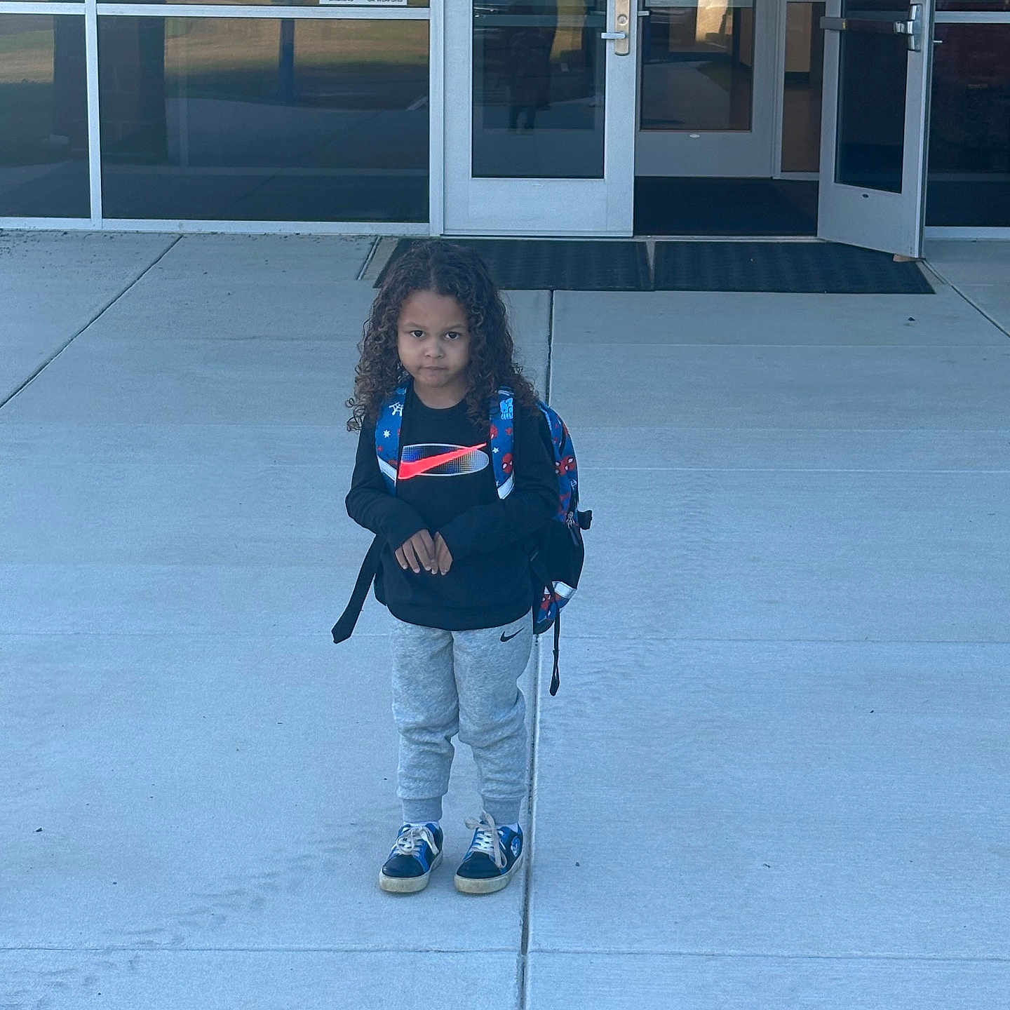Carsen joined the competition — help win amazing prizes! child, school, backpack, standing, outdoor, sidewalk, entrance, building, door, glass, curly_hair, portrait, full_body, clothing, sweatpants, sneakers, school_day, serious, person, architecture