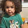 Carsen is registered to the contest to win money with this photo: child, toddler, curly_hair, portrait, indoors, green_shirt, graphic_tshirt, dog, dinosaur, serious_expression, face, person, sitting, chair, toy, window, wood_floor, home, natural_light, photography