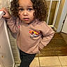 child, toddler, curly_hair, brown_hair, portrait, standing, indoor, bathroom, sweatshirt, race_car, black_pants, shoes, tile_floor, wood_floor, door, mirror, hand_on_hip, serious_expression, blue_eyes, casual_clothing