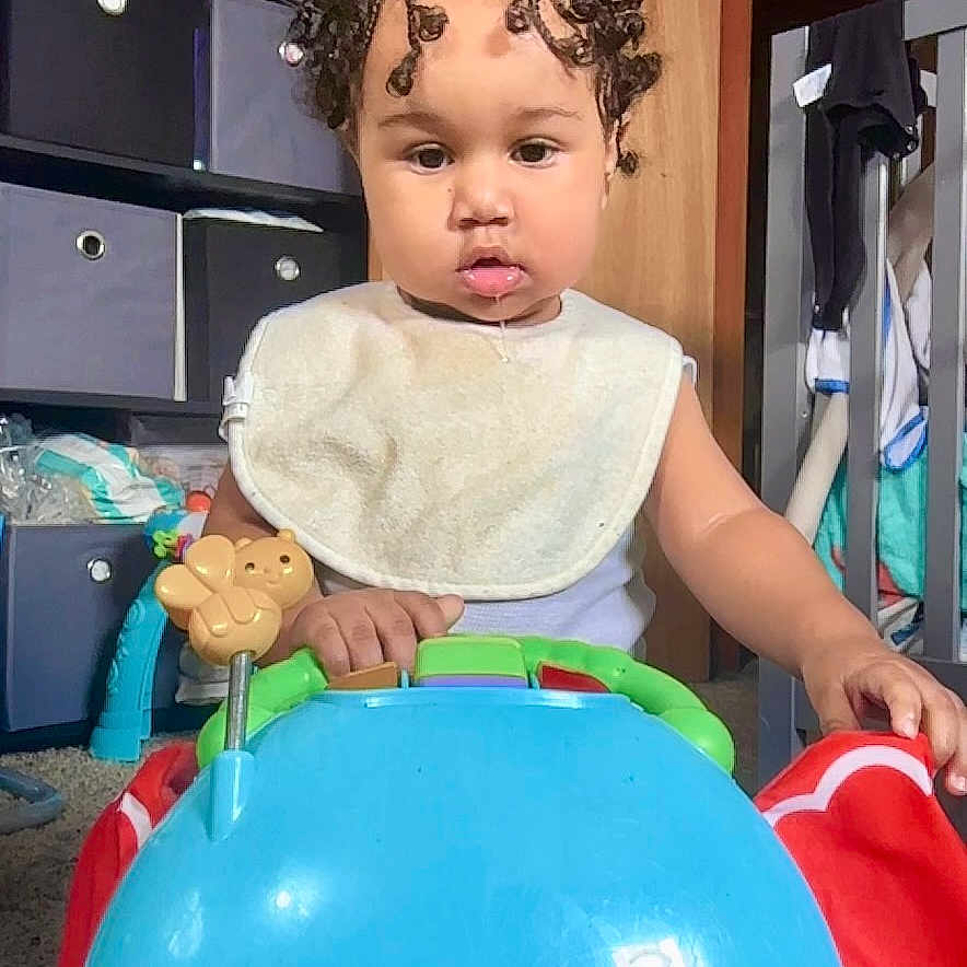 Wysdom is registered to the contest to win money with this photo: toddler, child, toy, elephant_toy, bib, curly_hair, indoor, baby, playtime, furniture, storage, cabinet, room, carpet, baby_gear, curiosity, person, young_child, cute, childhood