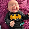 baby, infant, smile, laughing, batman_logo, hoodie, black_clothing, purple_blanket, texture, lying_down, indoors, cute, happy, child, pacifier_clip, hand, face, head, person, portrait