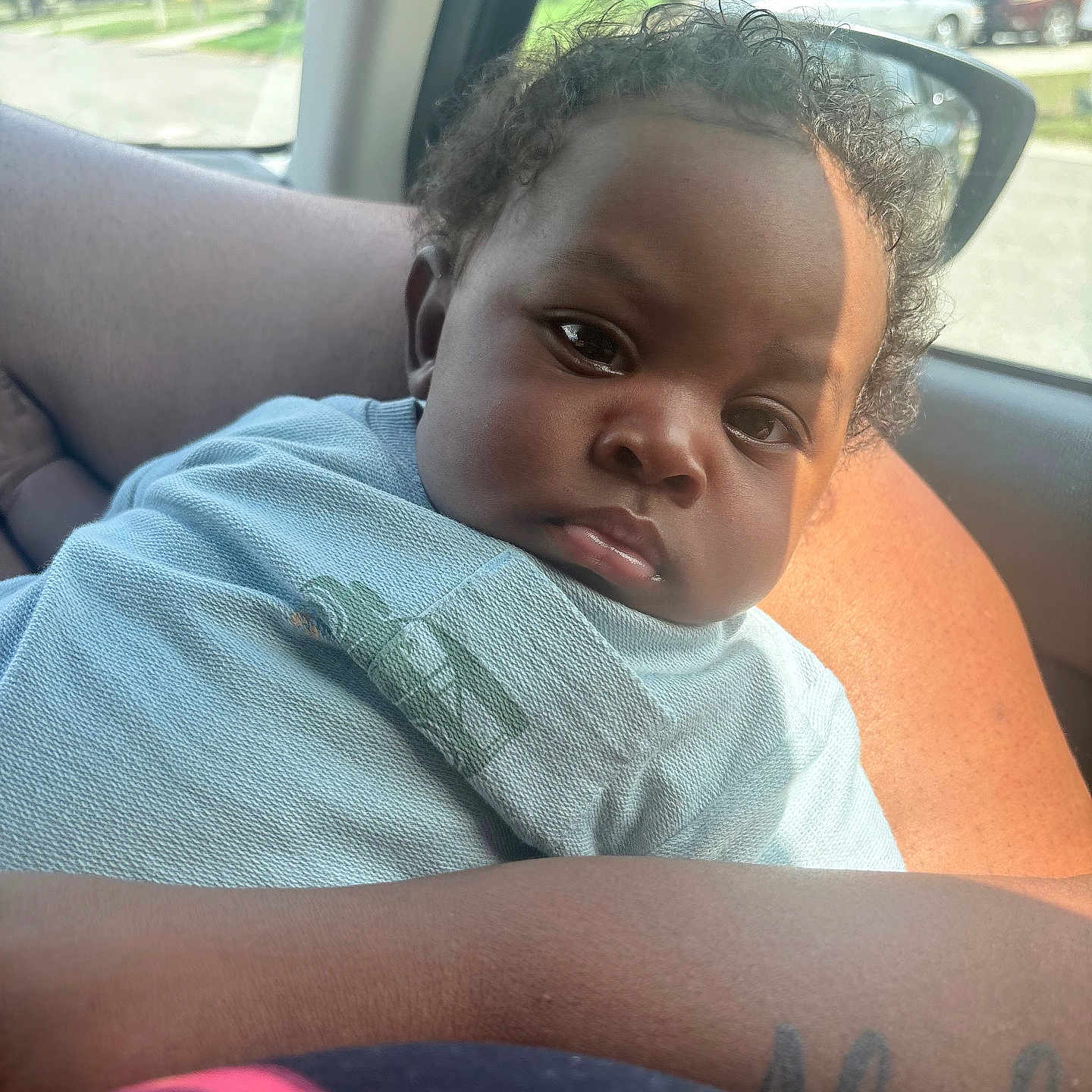 Kenzo is registered to the contest to win money with this photo: baby, child, curly_hair, car_interior, person, arm, blue_clothing, seatbelt, window, daylight, skin, portrait, cute, infant, holding, tattoo, side_view_mirror, expression, closeup, casual