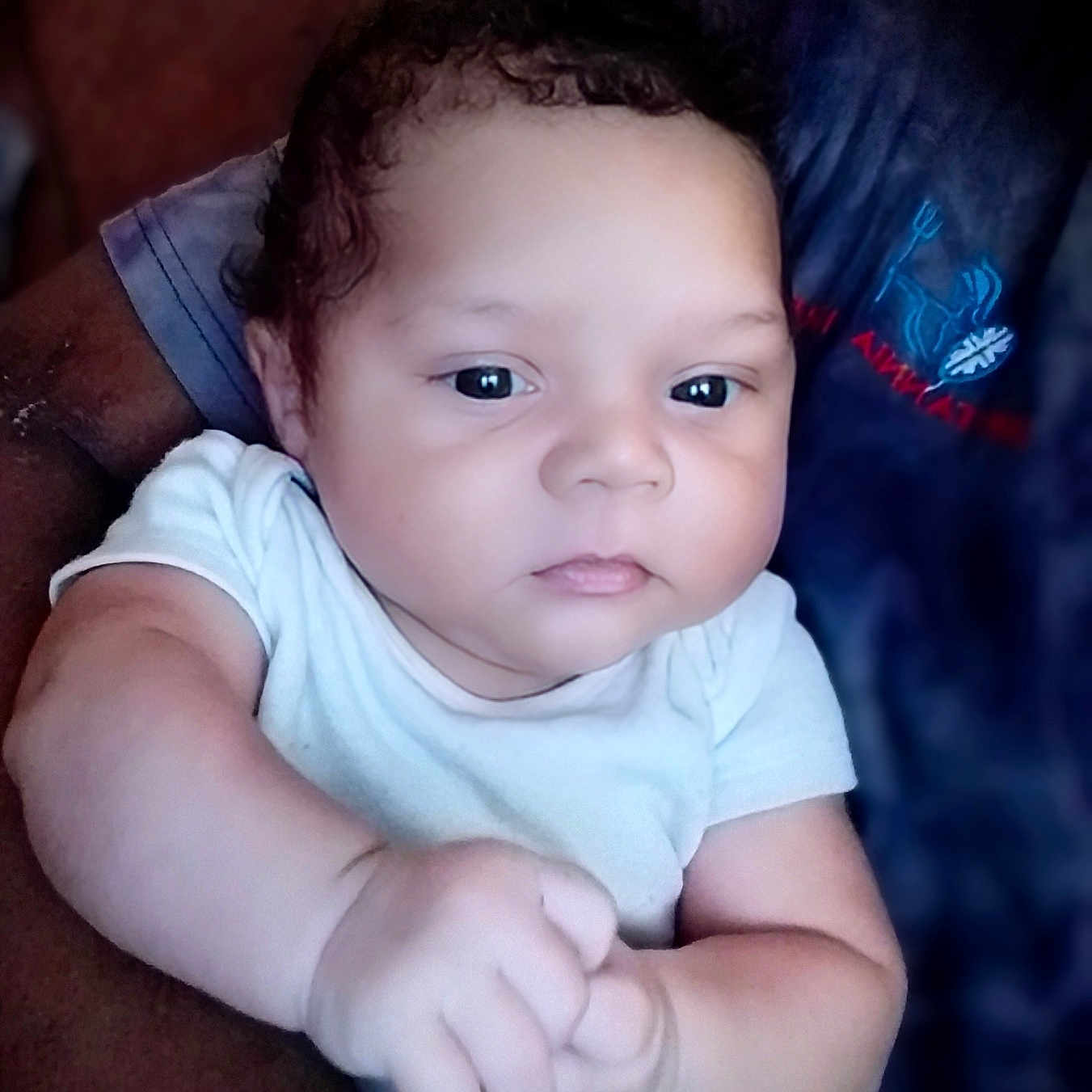 Grayson is registered to the contest to win money with this photo: adult_arm, arm, baby, casual, child, closeup, curly_hair, expressionless, face, hand, holding, indoor, infant, logo, person, portrait, seated, skin, tshirt, white_clothing