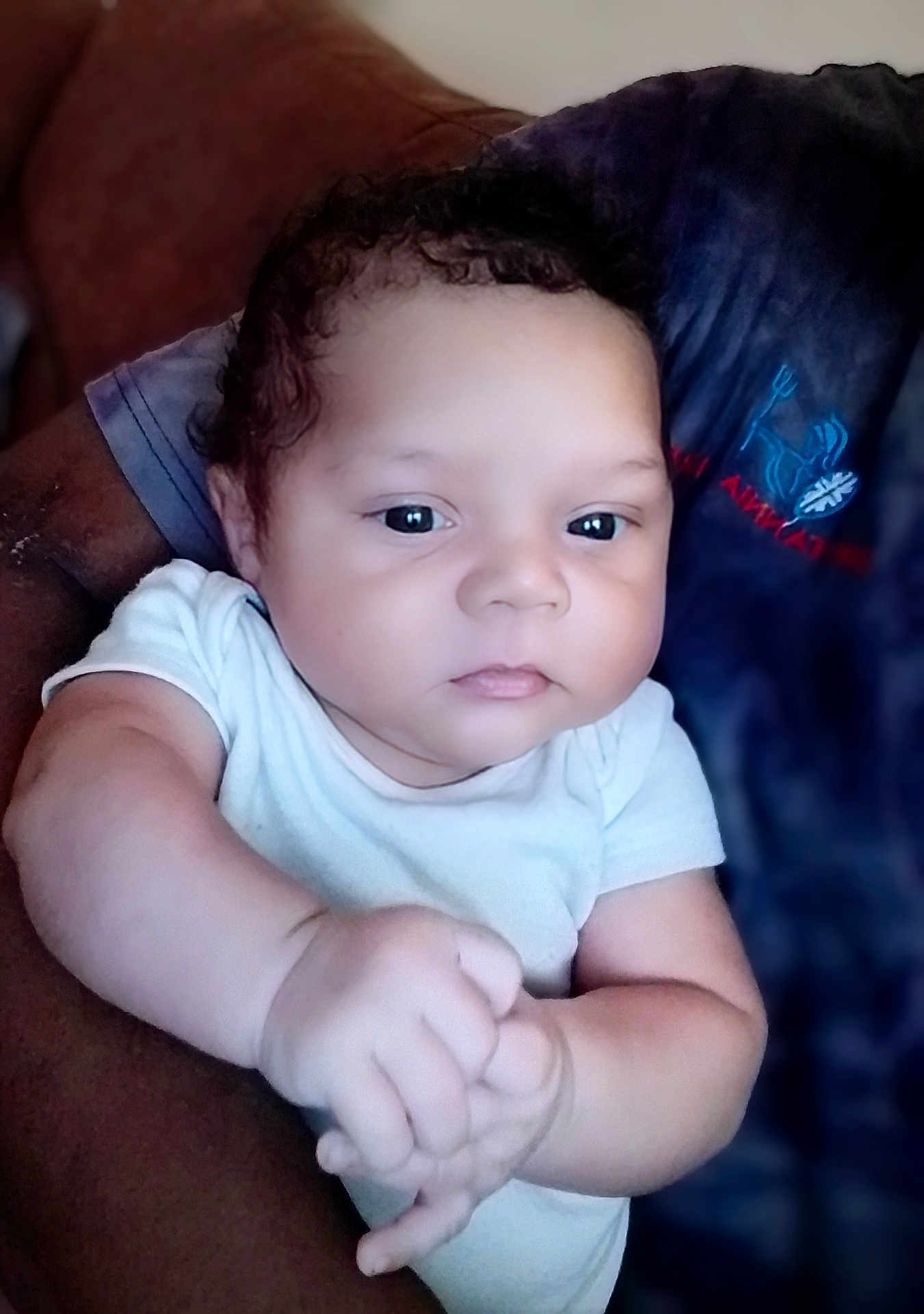 Grayson is registered to the contest to win money with this photo: baby, infant, child, person, arm, hand, face, curly_hair, white_clothing, adult_arm, holding, indoor, portrait, closeup, skin, expressionless, casual, tshirt, logo, seated