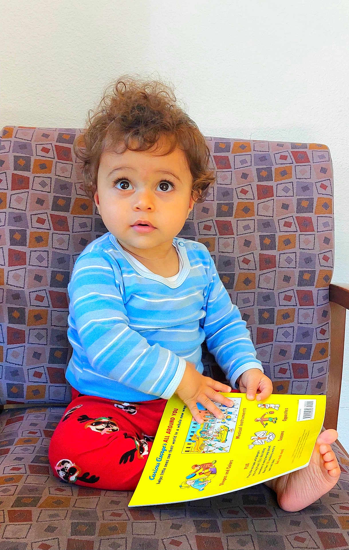 Antony is registered to the contest to win money with this photo: toddler, child, curly_hair, striped_shirt, red_pants, book, reading, chair, patterned_chair, barefoot, indoor, curiosity, wide_eyes, hands, foot, expression, young_child, casual_clothing, seated, portrait