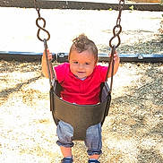 Antony is registered to the contest to win money with this photo: toddler, child, swing, playground, outdoor, sunlight, smiling, red_shirt, striped_pants, shoes, wood_chips, metal_chains, fence, play_area, baby, happy, young_child, casual_clothing, daytime, park