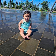 Francesca is registered to the contest to win money with this photo: toddler, child, swimsuit, water, splash_pad, outdoor, tiles, wet_surface, sunny, trees, fountain, playground, summer, park, sitting, person, head, face, barefoot, daytime