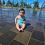 toddler, child, swimsuit, water, splash_pad, outdoor, tiles, wet_surface, sunny, trees, fountain, playground, summer, park, sitting, person, head, face, barefoot, daytime