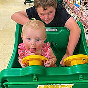 Raylynn is registered to the contest to win money with this photo: baby, child, shopping_cart, store, grocery, playful, tongue_out, pink_clothing, yellow_steering_wheel, green_cart, aisle, shelves, snacks, floor, person, fun, indoor, cute, two_children, supermarket
