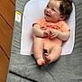 baby, infant, smiling, happy, curly_hair, onesie, baby_seat, cushion, gray_fabric, indoor, wooden_floor, cute, child, person, relaxed, small_feet, chubby_arms, comfortable, portrait, casual