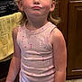 child, kid, smartphone, pink_tank_top, heart_pattern, kitchen, wooden_cabinet, yellow_towel, flooring, earrings, face, expression, standing, indoor, device, technology, casual_clothing, person, portrait, playful
