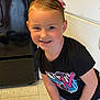child, smiling, hair_clip, black_tshirt, mtv_logo, kitchen, stove, floor, tile_floor, shorts, standing, happy, young_child, indoor, casual_clothing, person, portrait, blue_eyes, short_hair, light_skin