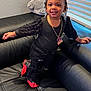 toddler, child, smiling, black_clothing, red_shoes, leather_couch, plush_toy, elephant_plush, indoors, window_blinds, hair_beads, necklace, cute, happy, standing, person, home, casual, portrait, fashion