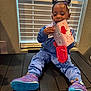 child, toddler, sitting, blue_tracksuit, purple_sneakers, hello_kitty, box, red_mitten, window_blinds, indoor, curtains, floor, toy, person, clothing, footwear, hair_buns, playful, smiling, young_child
