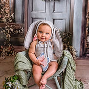 Remi is registered to the contest to win money with this photo: baby, child, bunny_ears, wicker_chair, butterfly_toy, flower_arrangement, green_fabric, portrait, indoor, cute, smiling, costume, toddler, decor, soft_lighting, seated, playful, infant, adorable, photography