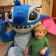 Garrett is registered to the contest to win money with this photo: child, mascot, blue, cartoon_character, indoor, classroom, smiling, green_shirt, blonde_hair, flooring, furniture, large_eyes, pink_ears, hand_holding, playful, costume, person, toy, happy, kid
