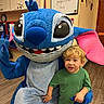 child, mascot, blue, cartoon_character, indoor, classroom, smiling, green_shirt, blonde_hair, flooring, furniture, large_eyes, pink_ears, hand_holding, playful, costume, person, toy, happy, kid
