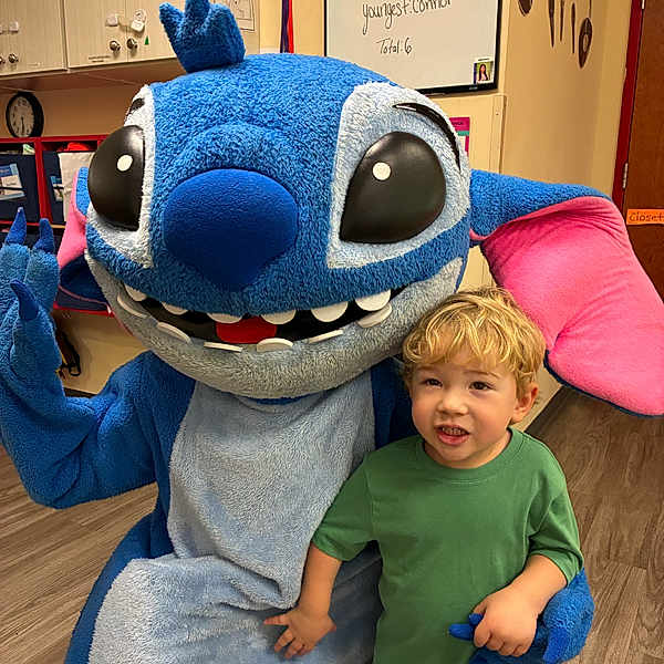 Garrett is registered to the contest to win money with this photo: child, mascot, blue, cartoon_character, indoor, classroom, smiling, green_shirt, blonde_hair, flooring, furniture, large_eyes, pink_ears, hand_holding, playful, costume, person, toy, happy, kid