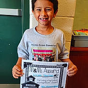 Elijah joined the competition — help win amazing prizes! child, boy, smiling, award, certificate, tshirt, school, indoor, brick_wall, green_door, missing_tooth, proud, holding, portrait, young, casual_clothing, class_photo, education, student, person