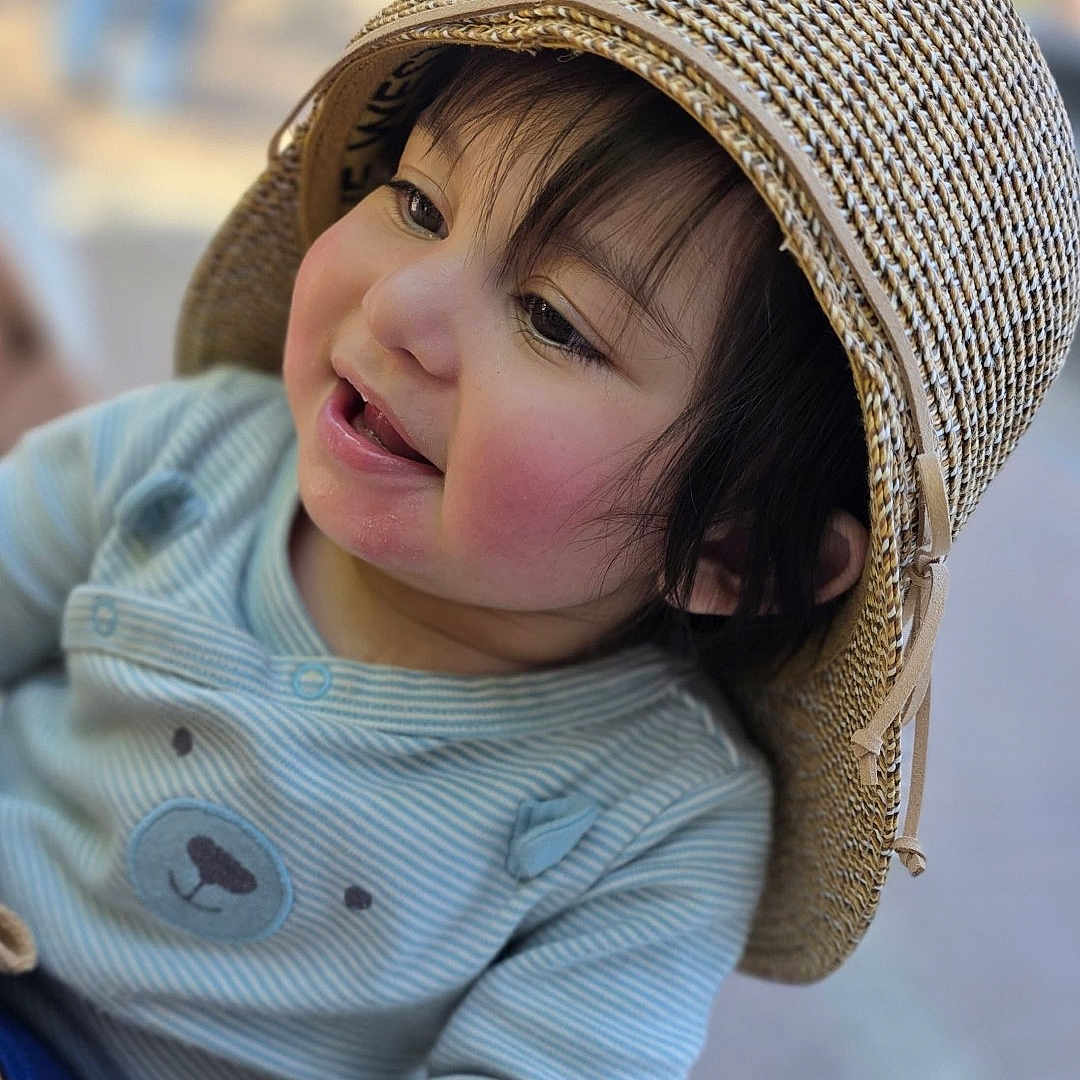 Eshaal is registered to the contest to win money with this photo: baby, blue_shirt, blurred_background, child, close_up, clothing, eyes, face, hair, happy, infant, joy, outdoor, photography, portrait, seated, smiling, sun_hat, toddler, woven_hat